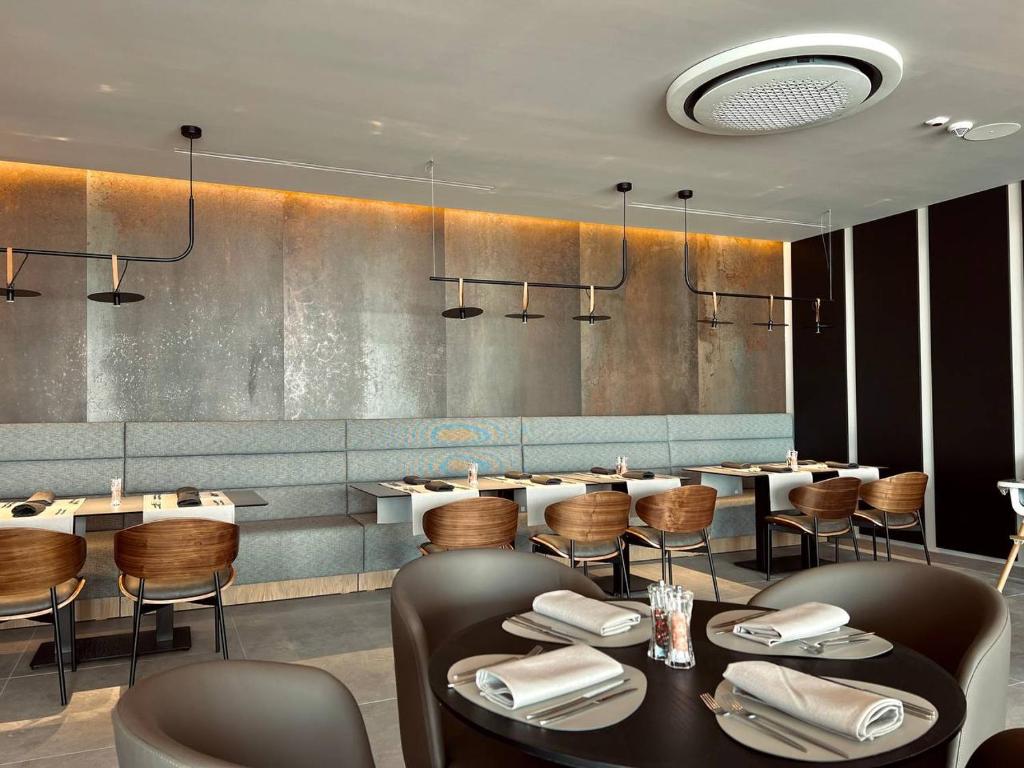 Mūsdienīgs restorāns ar elegantu interjeru, kurā ir stilīgas koka mēbeles un atmosfēriska apgaismojuma elementi. Galdi ir sakārtoti ar uzklātiem galda piederumiem un salvetēm.