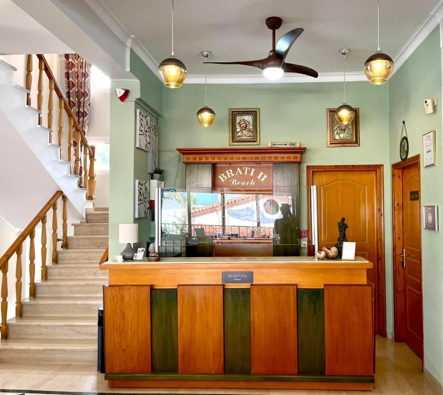 Recepcija viesnīcā ar elegantām detaļām un modernu dizainu, tajā ir koka galds ar uzrakstu "BRATI II Beach" un mākslas darbi uz sienām. Gaismas elementi un kāpnes piešķir telpai viesmīlīgu atmosfēru.