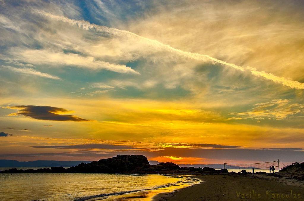 Saulriets pie pludmales ar krāsainām debesīm un mierīgu jūru, kur redzami cilvēki, kas staigā pa krastu.