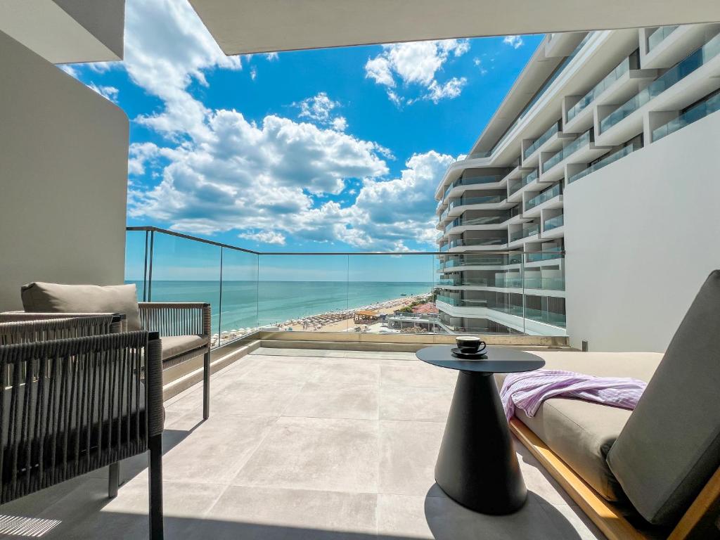 Mūsdienīgs balkons ar skatu uz jūru un pludmali, kurā ir ērtas mēbeles un glīta kafijas tasīte uz galda. Debesis ir skaidras un saulainas, radot relaksējošu atmosfēru.