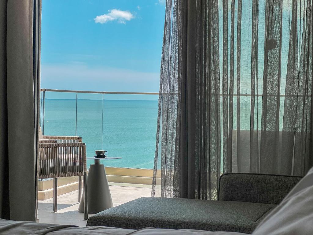 Skats no viesnīcas numura uz jūras krastu, caur caurspīdīgu aizkaru. Uz balkona redzama kafejnīca ar krūzi un mūsdienīgu mēbeļu dizainu.