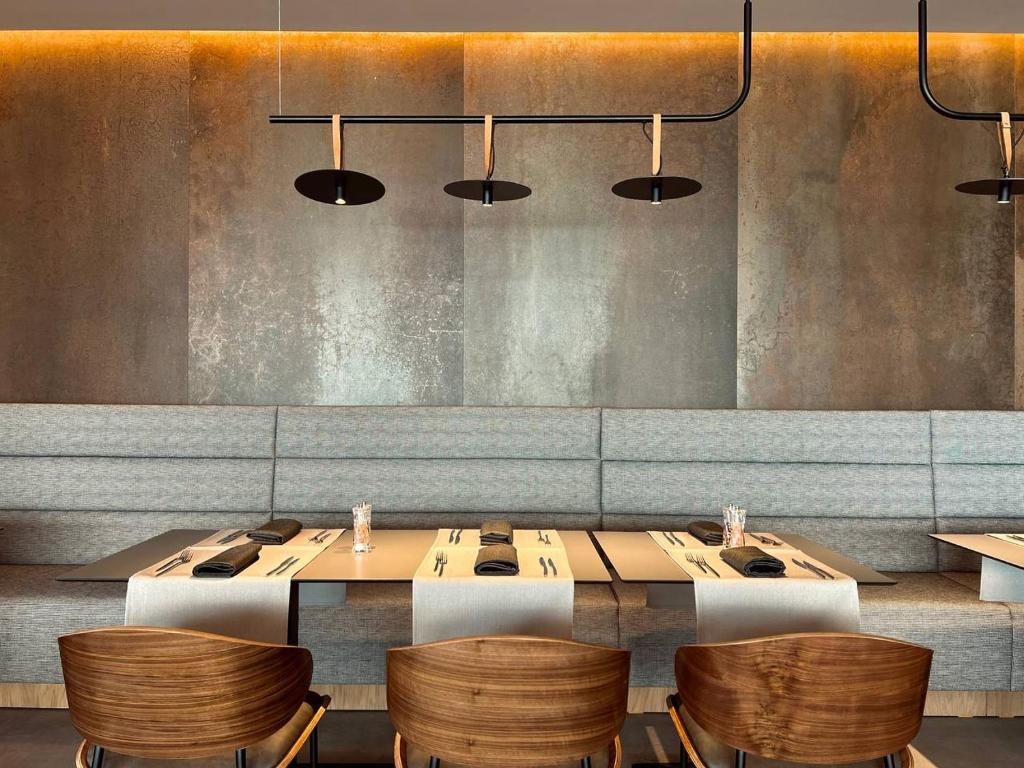 Mūsdienīga restorāna interjers ar pelēku sēdvietu un koka krēsliem, kas izgaismots ar stilīgām lampām. Galdi ir sakārtoti ar galda piederumiem un salvetēm, radot elegantu atmosfēru.