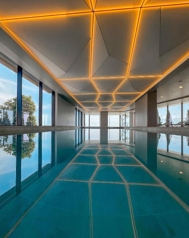 Mūsdienīgs peldbaseins ar ģeometrisku griestu apgaismojumu un lieliem logiem, caur kuriem ieplūst dabiska gaisma. Ūdens virsma atspoguļo telpas dizainu un apkārtējo dabu.
