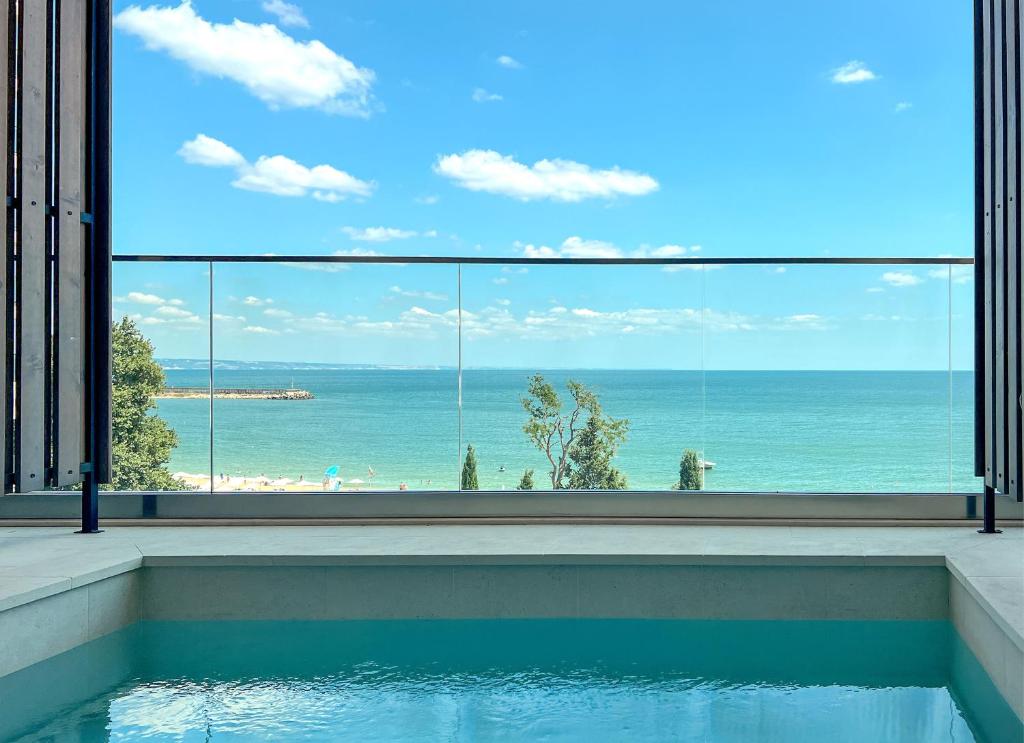 Skats uz jūru no modernas terases ar stikla barjeru, kurā redzams baseins un zila debess ar baltām mākoņiem.