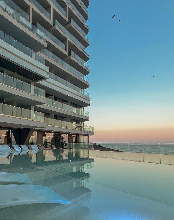 Mūsdienīga ēka ar stikla balkoniem un baseinu, kas atspoguļo saulrietu pie jūras. Skats uz debesīm ar viegliem mākoņiem un putniem lidojot.