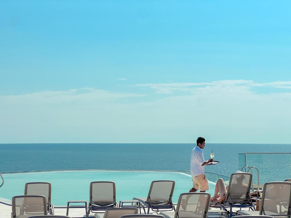 Viesmīlis nes glāzi uz baseina malas, kamēr viesi relaksējas pie ūdens. Skaists skats uz jūru un debesi rada mierīgu atmosfēru.