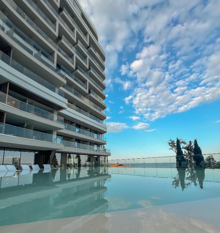 Mūsdienīgs dzīvokļu komplekss ar stikla fasādi, kas atspoguļojas baseinā zem skaidra, zila debesu fona.