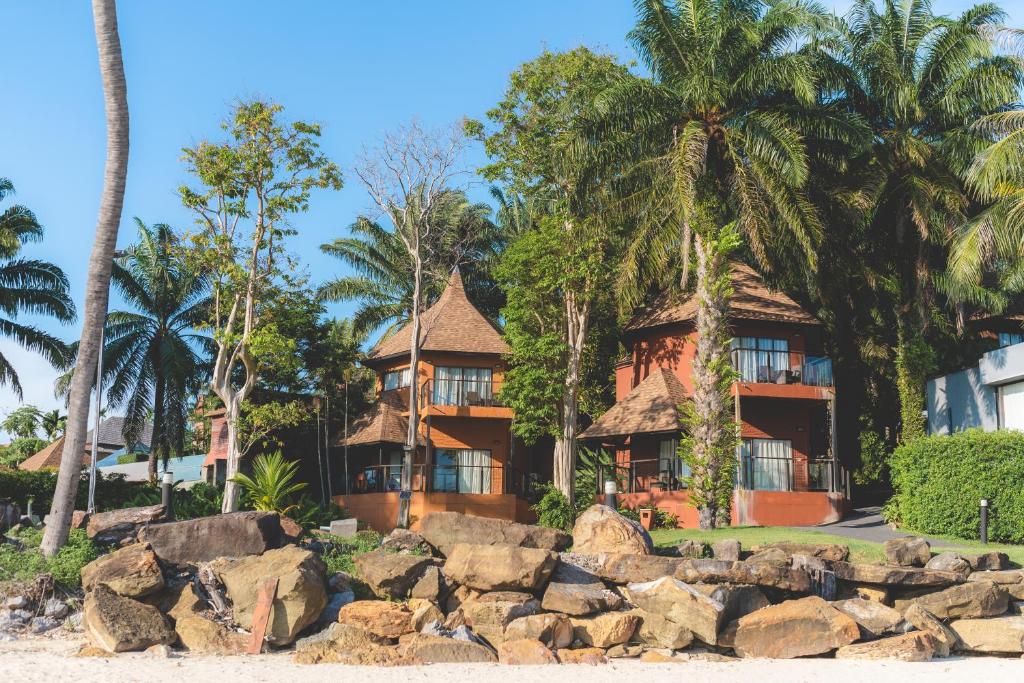 Tropu ainava ar stilīgām mājām un palmām, kas atrodas pie pludmales, akmeņi priekšplānā.