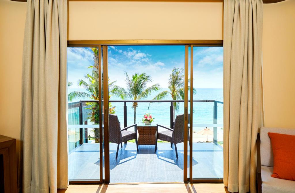 Skats uz jūru no viesnīcas numura, kurā redzami divi ērtie krēsli uz balkona, blakus palmām un ziediem. Gaisma plūst caur plašajiem logiem, radot relaksējošu atmosfēru.