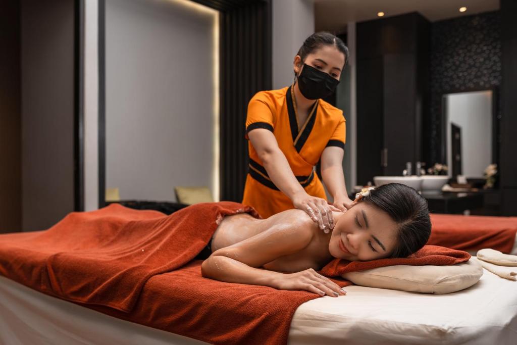 Masāžas sesija spa centrā, kur masieris rūpīgi apstrādā klienta muguru. Klients relaksējas uz masāžas galda ar sarkanu dvieli un ziedu matos.