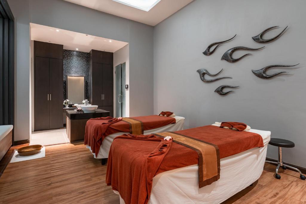 Mūsdienīgs spa telpas interjers ar divām masāžas gultām, siltām toņu segām un dekoratīviem elementiem uz sienas. Telpā ir arī eleganta izlietne un spogulis, radot relaksējošu atmosfēru.