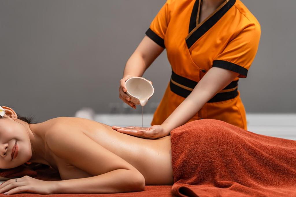 Masāžas sesija, kur masieris uzklāj eļļu uz klienta muguras, radot relaksējošu atmosfēru. Klients atpūšas uz masāžas galda, pārklāts ar dvieli.