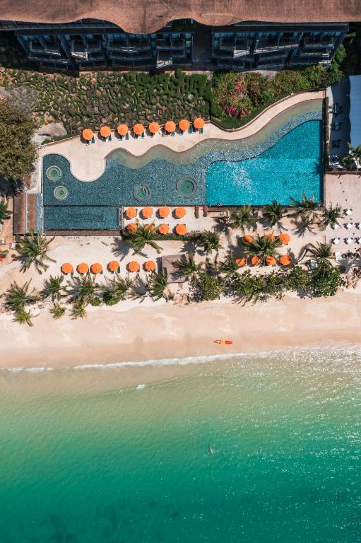 Tuvplāns ar baseinu un pludmali, kurā ir oranži saulessargi un palmām. Ūdens ir dzidrs, un pludmale ir smilšaina, piedāvājot relaksējošu atmosfēru.