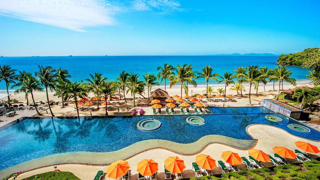 Viesnīcas baseins ar oranžām saules parasolēm un palmu kokiem, blakus skaistai pludmalei un jūrai. Ideāla vieta atpūtai un relaksācijai.