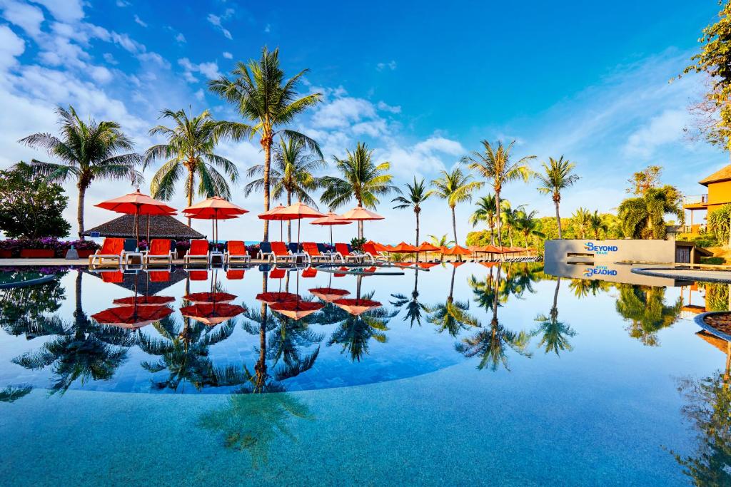 Tropu kūrorta baseins ar sarkaniem saulessargiem, palmām un skaidru zilu debesu fona, atspoguļojot mierīgu atmosfēru.