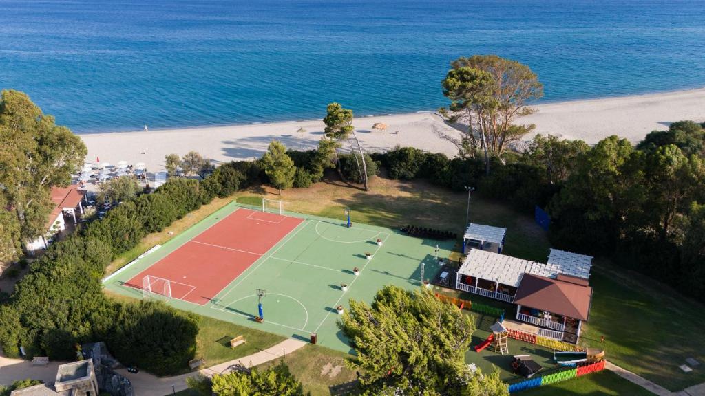Skaists skats uz pludmales sporta laukumiem, kas atrodas pie jūras, ar zaļu zāli un sarkanu laukumu. Fons veido mierīga pludmale un zila jūra, radot relaksējošu atmosfēru.