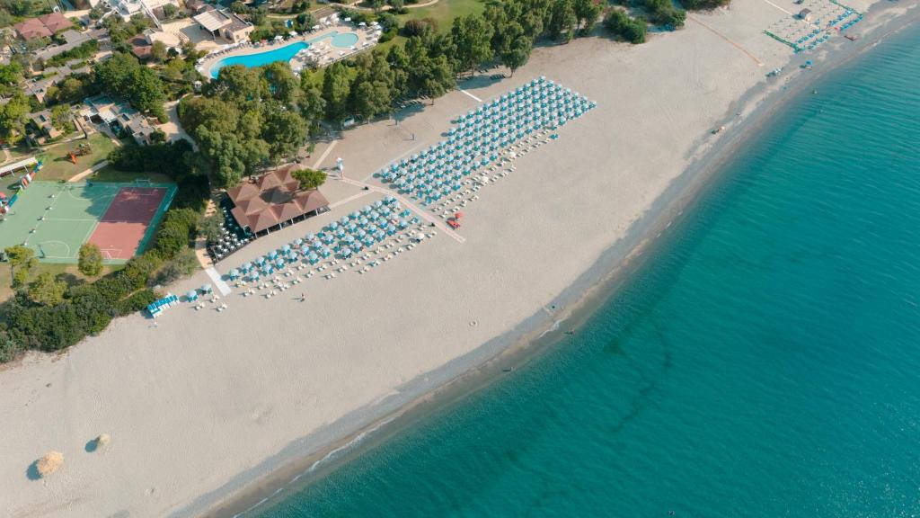 Plaša pludmale ar saulessargiem, skats no augšas, blakus viesnīcai un peldbaseinam. Zaļš fons ar kokiem un sporta laukumiem.