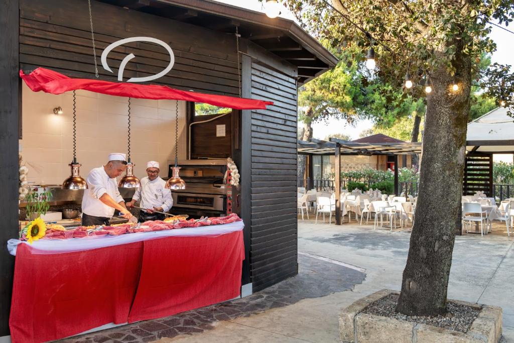 Āra ēstuve ar šefpavāriem, kas gatavo ēdienus, un viesmīlīgu atmosfēru, apvienojot dabu un gastronomiju. Redzami galdi un krēsli, kas gaida viesus.