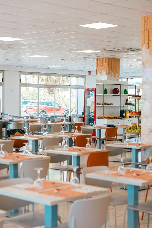 Mūsdienīgs restorāna interjers ar gaišām krāsām, koka elementiem un lieliem logiem, kas ļauj iekļūt dabiskajai gaismai. Uz galdiem ir salvetes un glāzes, radot viesmīlīgu atmosfēru.