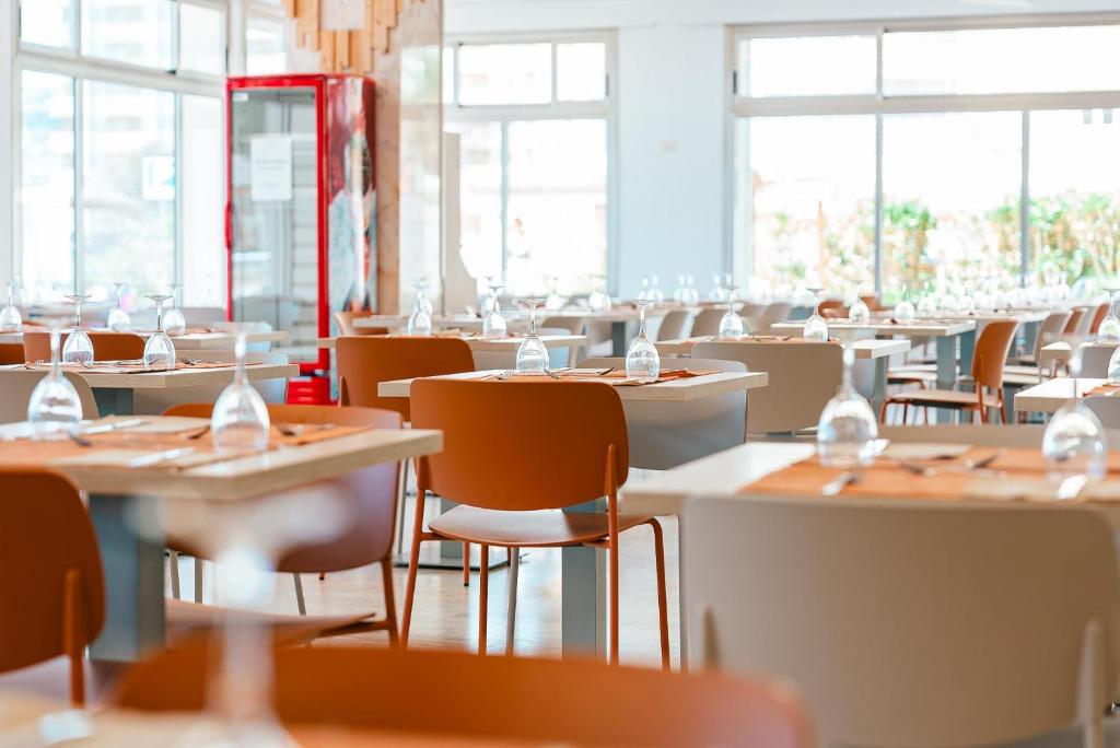 Mūsdienīgs restorāns ar stilīgiem galdiem un krēsliem, kas izvietoti gaišā, gaisīgā telpā. Redzamas glāzes un galda piederumi, kas liecina par rūpīgu sagatavošanu.