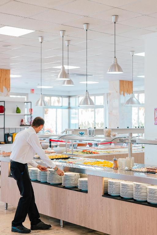 Pakalpojumu darbinieks sakārto traukus bufetē, kas piepildīta ar dažādiem ēdieniem, modernā un gaišā restorāna vidē.