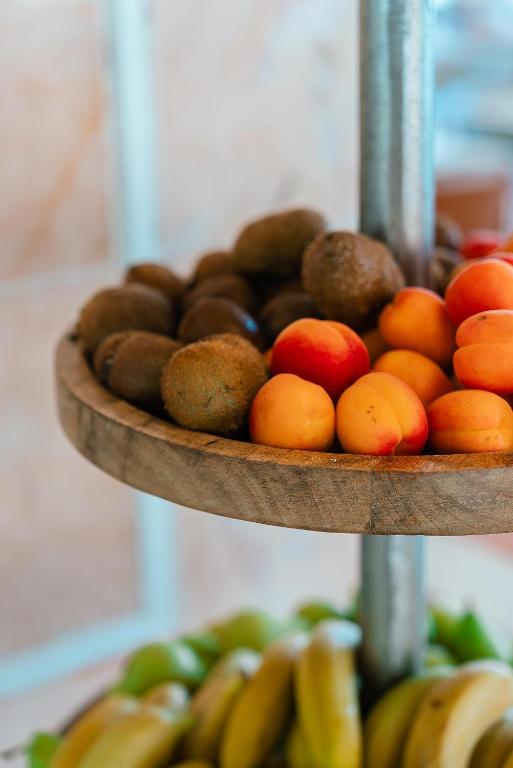 Augļu grozs ar kivi un aprikozēm, kas novietots uz koka statīva. Apakšā redzamas arī banānu šķirnes.