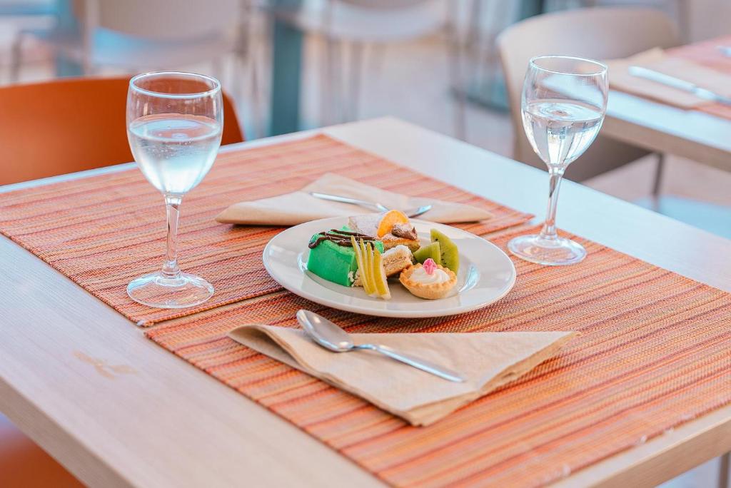 Galda uzstādījums ar divām glāzēm ūdens un desertu, kas ietver krāsainus saldumus un augļus, uz oranžas salvetes.