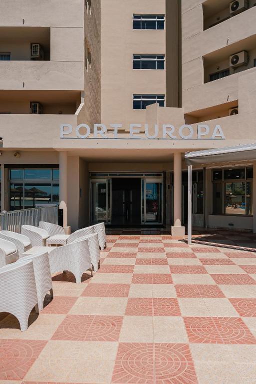 Viesnīcas "Port Europa" ieeja ar modernu arhitektūru un plašu terasi, kurā redzami baltie krēsli un flīžu zeme.