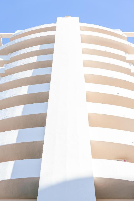 Mūsdienīga daudzstāvu ēka ar baltiem balkoniem un skaidru zilo debesu fonu. Architektūras detaļas izceļ eleganci un modernitāti.