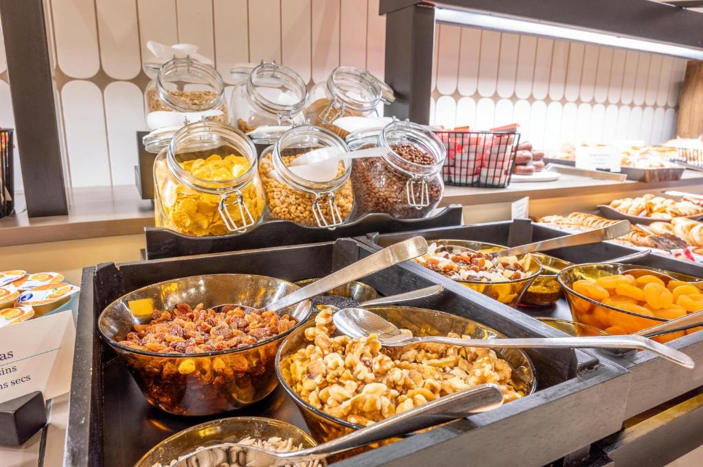 Pakalpojumu stendā redzami dažādi uzkodu veidi, tostarp rieksti, žāvēti augļi un graudaugi, kas pasniegti caurspīdīgās burkās un bļodās. Uzkodas ir sakārtotas pievilcīgi, radot viesmīlīgu atmosfēru.