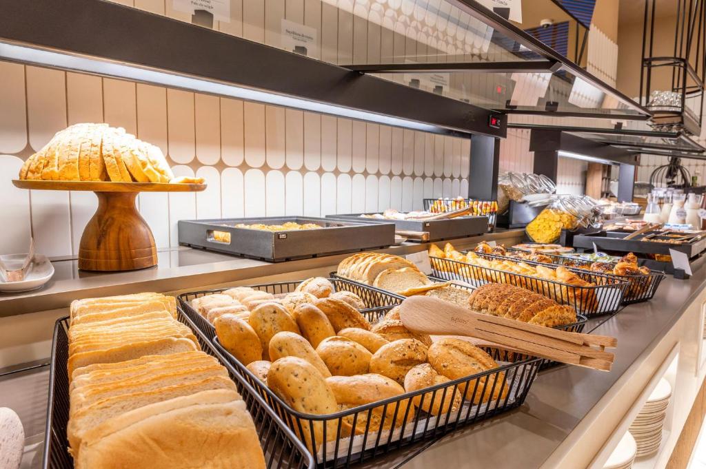 Šī ir brokastu bufete ar dažādiem maizes izstrādājumiem, tostarp šķēlēs sagrieztu maizi un bulciņām, kas pasniegtas stilīgās grozās. Aiz bufetes redzama modernā interjera dizains.
