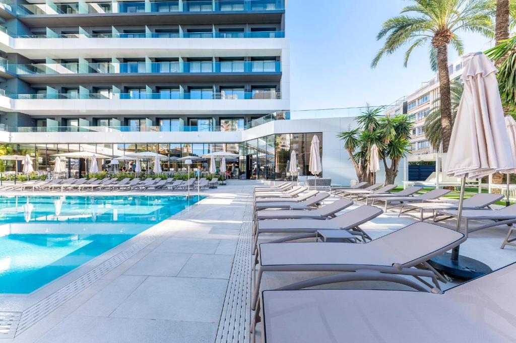 Mūsdienīgs viesnīcas baseina apgabals ar sauļošanās krēsliem un palmām, kas nodrošina relaksējošu atmosfēru. Labi pārskatāms skats uz modernu ēku un plašu terasi.