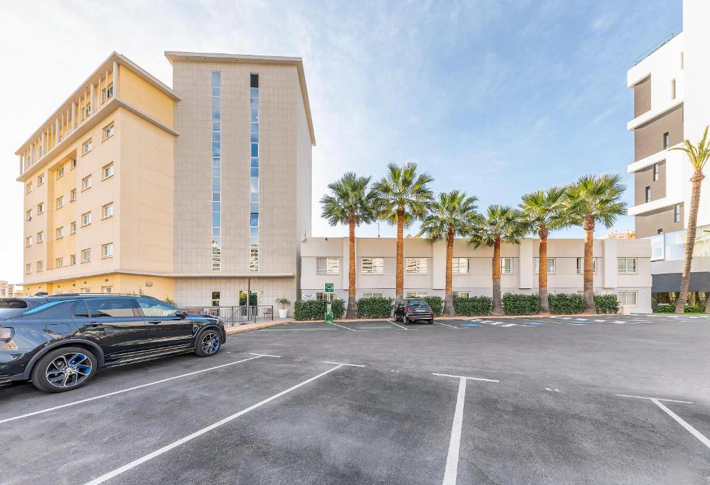 Mūsdienīga viesnīca ar palmām un autostāvvietu, kas atrodas saulainā apkārtnē. Redzami divi dažādi ēku stili un skaidrs debess fons.