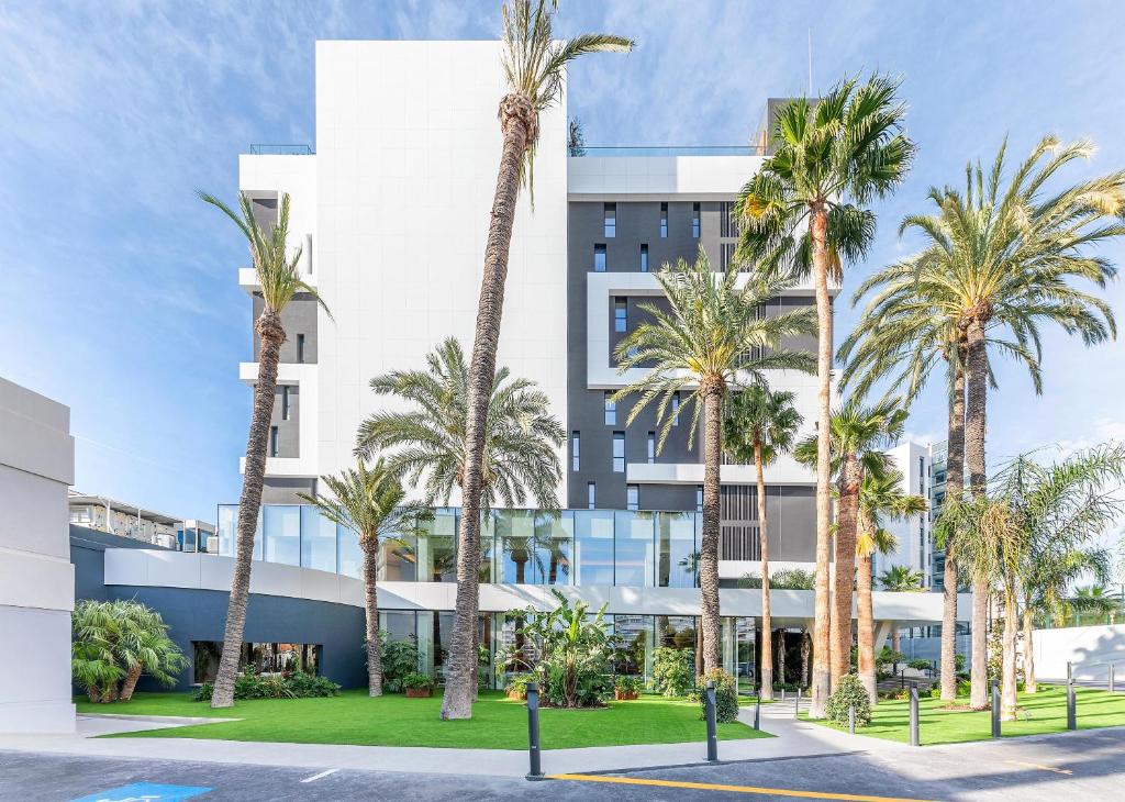 Mūsdienīga viesnīca ar plašu fasādi, ko ieskauj palmām un zaļš zāliena laukums, radot tropisku atmosfēru.