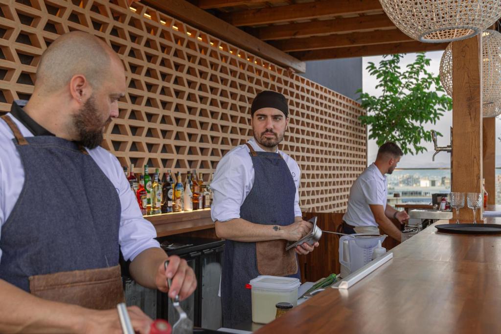 Šajā attēlā redzami trīs pavāri, kas strādā stilīgā bārā ar modernu dizainu. Viņi sagatavo dzērienus un ēdienus, kamēr fonā ir plaša dzērienu izvēle.