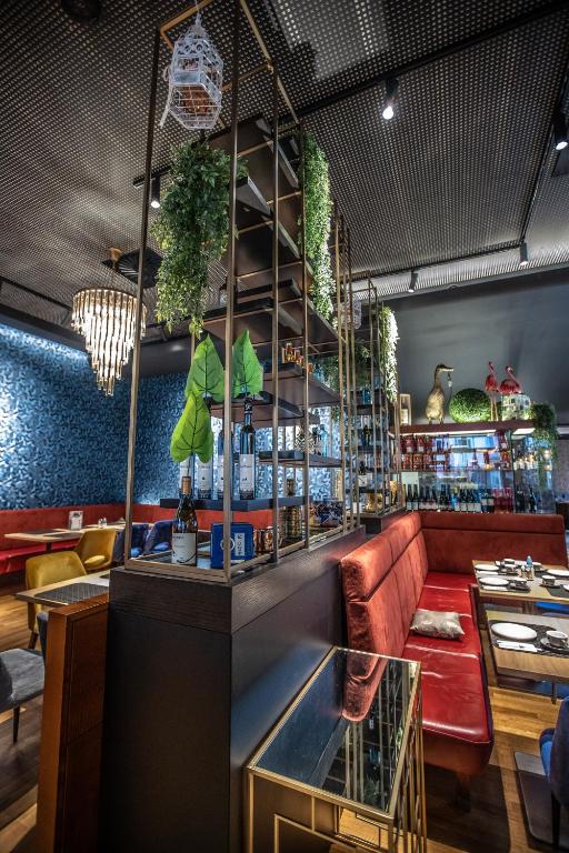 Mūsdienīga restorāna interjers ar elegantu mēbeļu izkārtojumu, dekoratīviem augiem un modernām gaismām. Sarkanās un dzeltenās krāsas akcenti piešķir dzīvīgumu telpai.