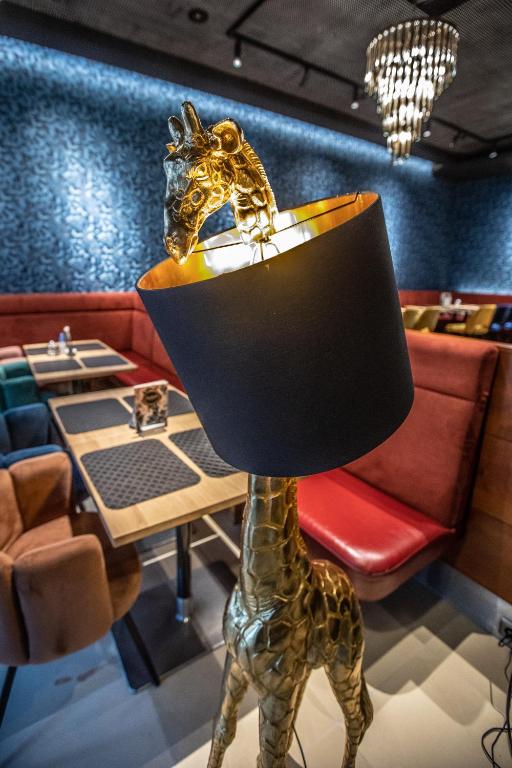 Zelta žirafes lampa ar melnu abažūru, kas atrodas stilīgā restorānā ar krāsainām mēbelēm un modernu interjeru. Izsmalcināta atmosfēra un radošs dizains.
