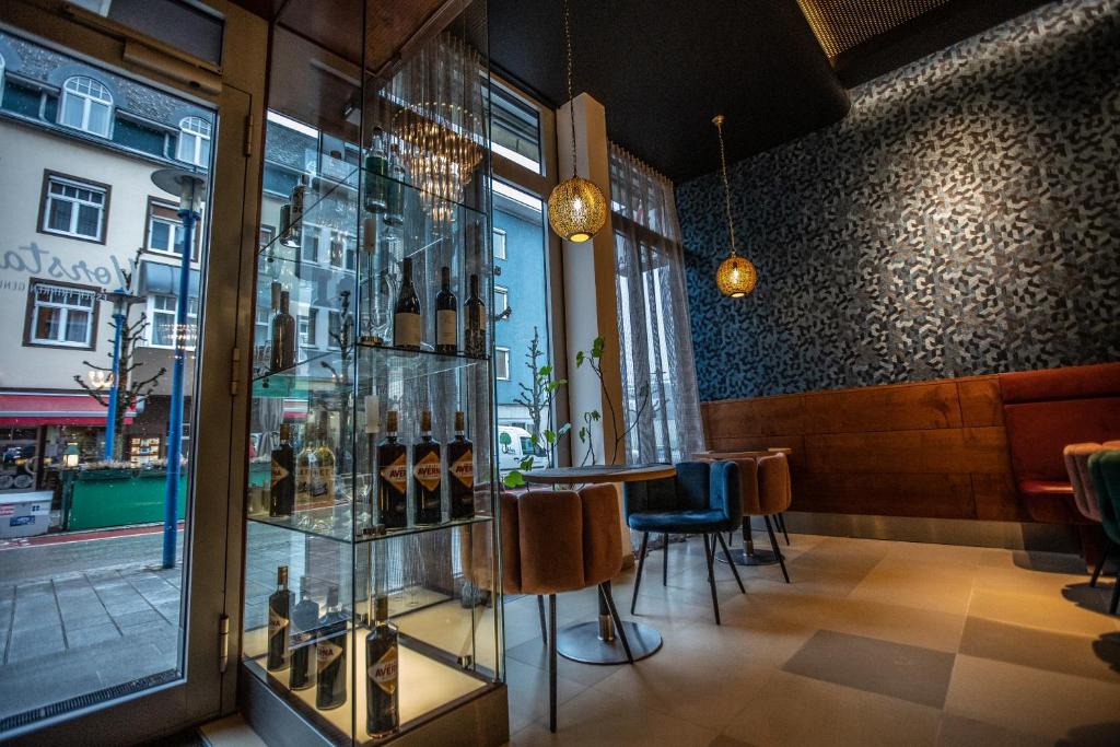 Mūsdienīgs restorāna interjers ar glītu dizainu, kurā redzama stikla plaukts ar vīna pudelēm un eleganti gaismas ķermeņi. Logu tuvumā atrodas krāsainas krēsli un galdi, radot viesmīlīgu atmosfēru.