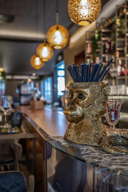 Zelta pērtiķa galvas dekorācija ar salmiņiem, kas atrodas bārā, radot unikālu un stilīgu atmosfēru. Aiz tā redzama gaiša bāra vide ar dekoratīvām lampām.
