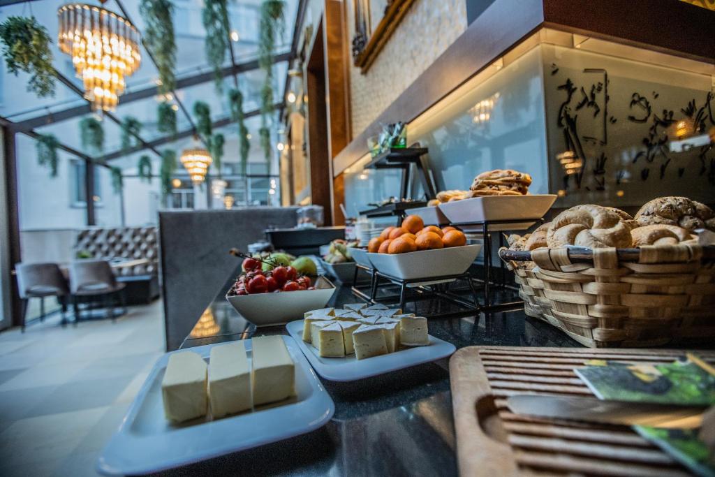 Bagātīgs brokastu galds ar dažādiem augļiem, maizēm un sieriem, viss elegantā restorāna vidē ar lieliem logiem un greznām lampām.