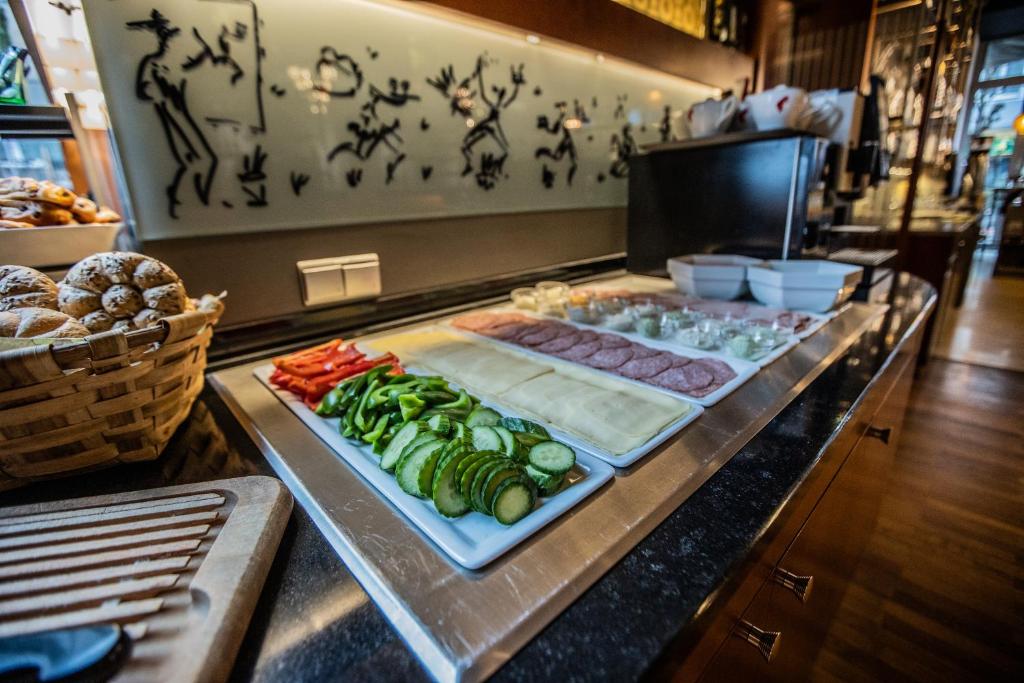 Pusdienu galda ar dažādām sastāvdaļām, tostarp dārzeņiem, gaļu un sieru, kā arī maizēm grozā. Fons ar māksliniecisku sienas dekorāciju.