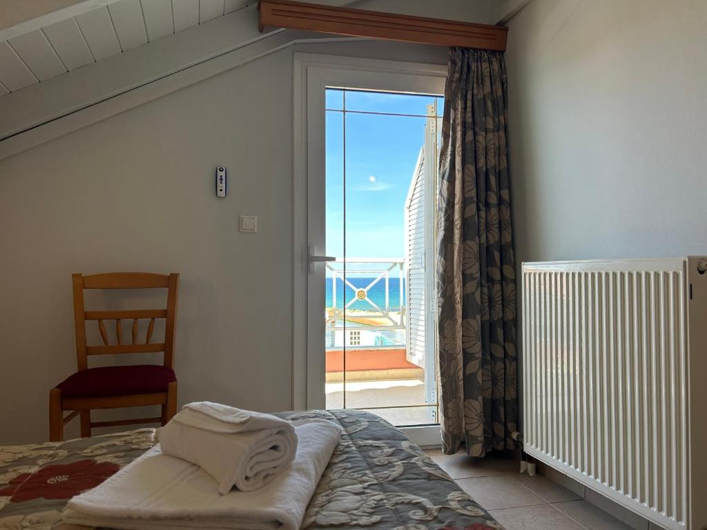 Gultas skats ar skatu uz jūru, kur redzama atvērta durvju loga ar aizkariem un koka krēsls telpā. Gultas veļa ar rakstu un salocītām dvieļiem uz gultas.