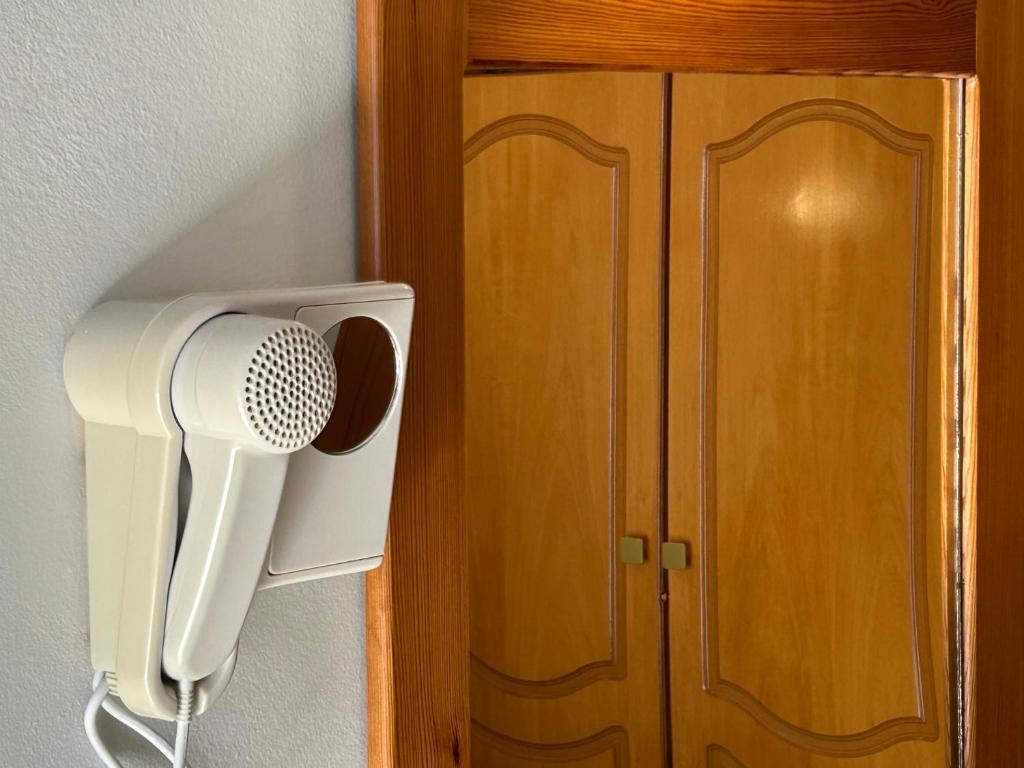 Sienā piestiprināts tālrunis ar mikrofonu, blakus koka durvīm. Tālrunis ir gaiši krāsots un atrodas telpā ar gaišu apgaismojumu.