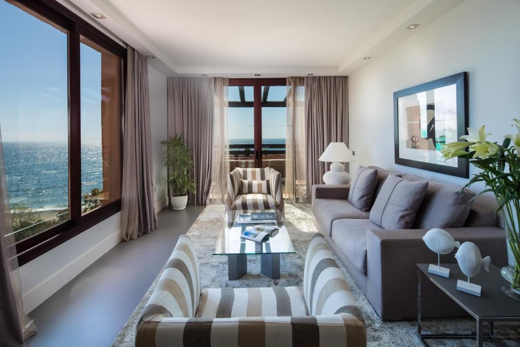 Mūsdienīga un eleganta viesistaba ar lieliem logiem, kas sniedz skatu uz jūru. Interjerā ir gaiši toņi, komfortabili dīvāni un dekoratīvi elementi, kas rada mierīgu atmosfēru.