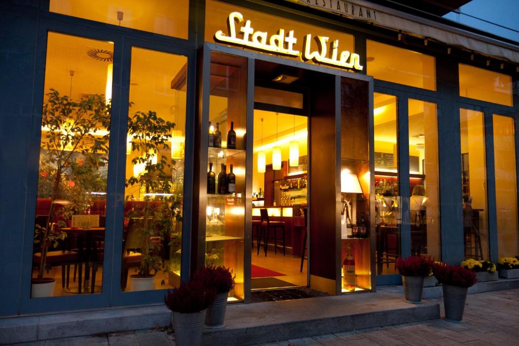 Restorāna "Stadt Wien" ieeja ar siltu apgaismojumu un elegantu interjeru, kas aicina apmeklētājus. Redzami koki un puķu podi pie durvīm.
