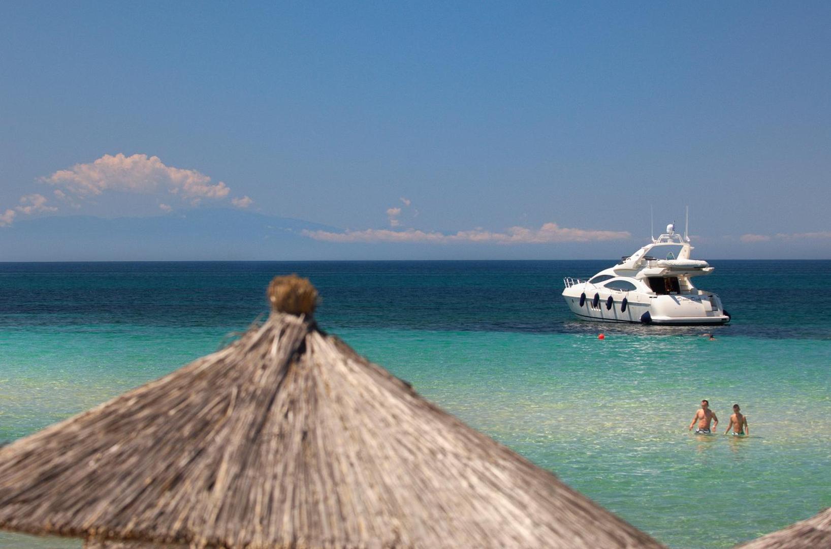 Portes Beach Hotel skats uz tirkīzzilo Egejas jūru Halkidiki piekrastē ar baltu jahtu un salmu saulessargu priekšplānā
