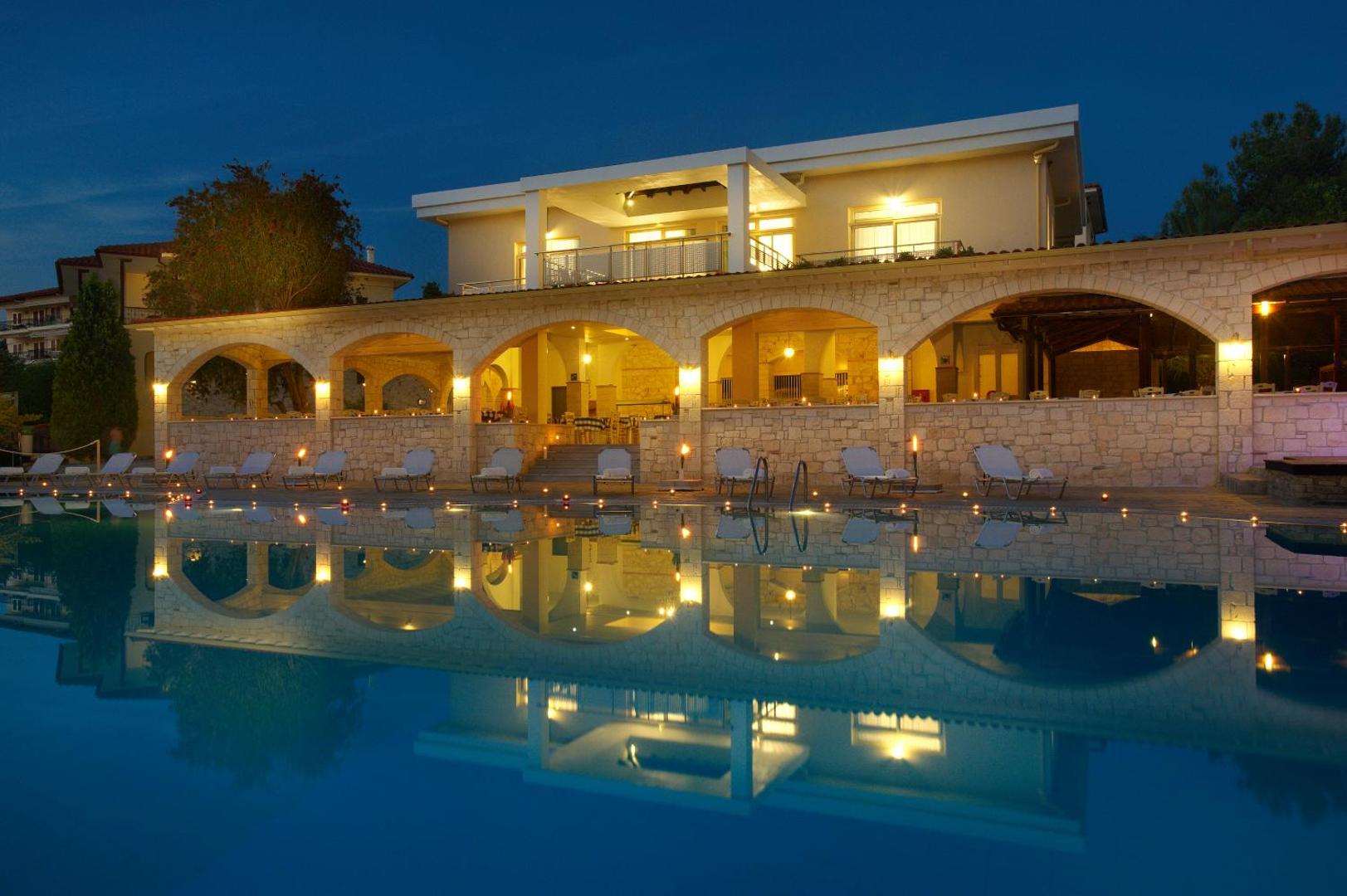 Portes Beach Hotel galvenā ēka un āra baseins vakarā Halkidiki piekrastē ar izgaismotām akmens arkām un atpūtas krēsliem pie ūdens