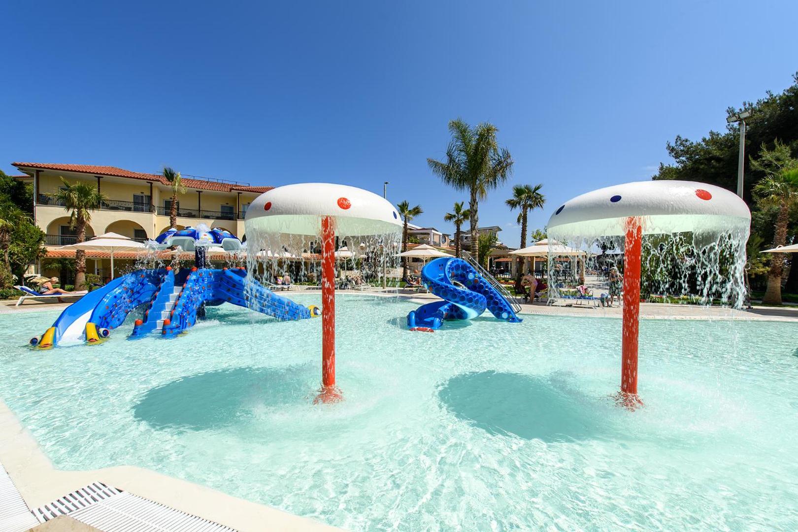 Portes Beach Hotel bērnu baseins Halkidiki piekrastē ar ziliem slidkalniņiem un ūdens sēnēm saulainā dienā, apkārt palmas un atpūtas krēsli