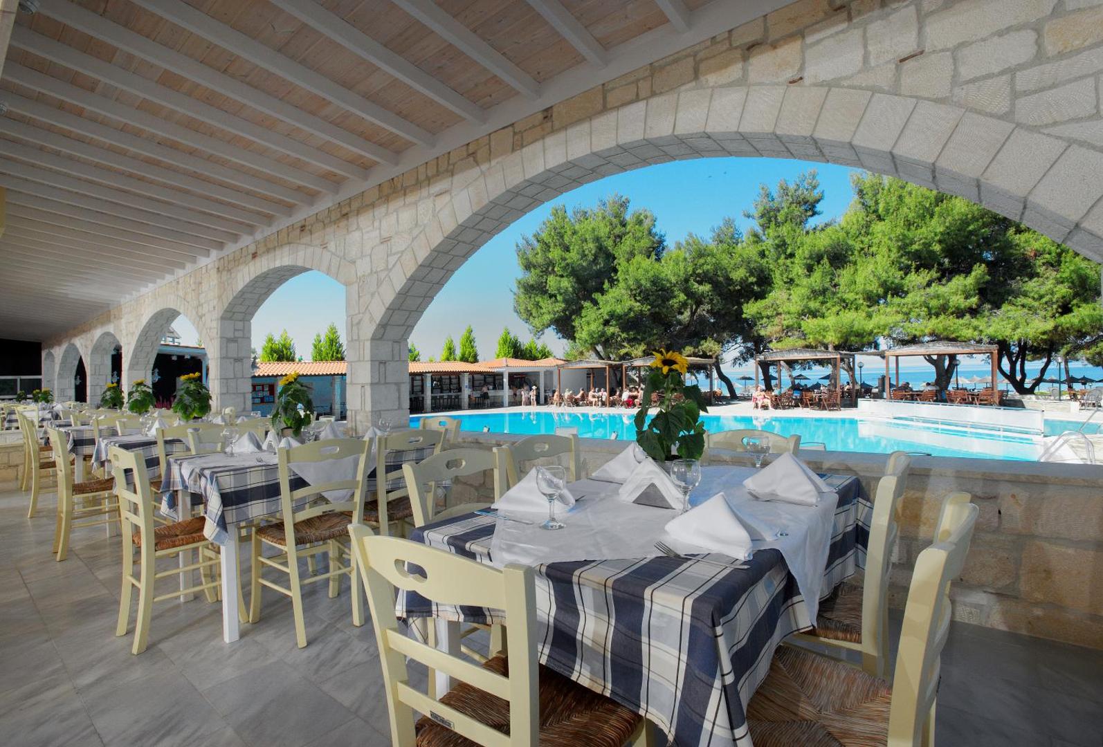 Portes Beach Hotel restorāna āra terase ar klātiem galdiem zem akmens arkām un skatu uz baseinu un dārzu