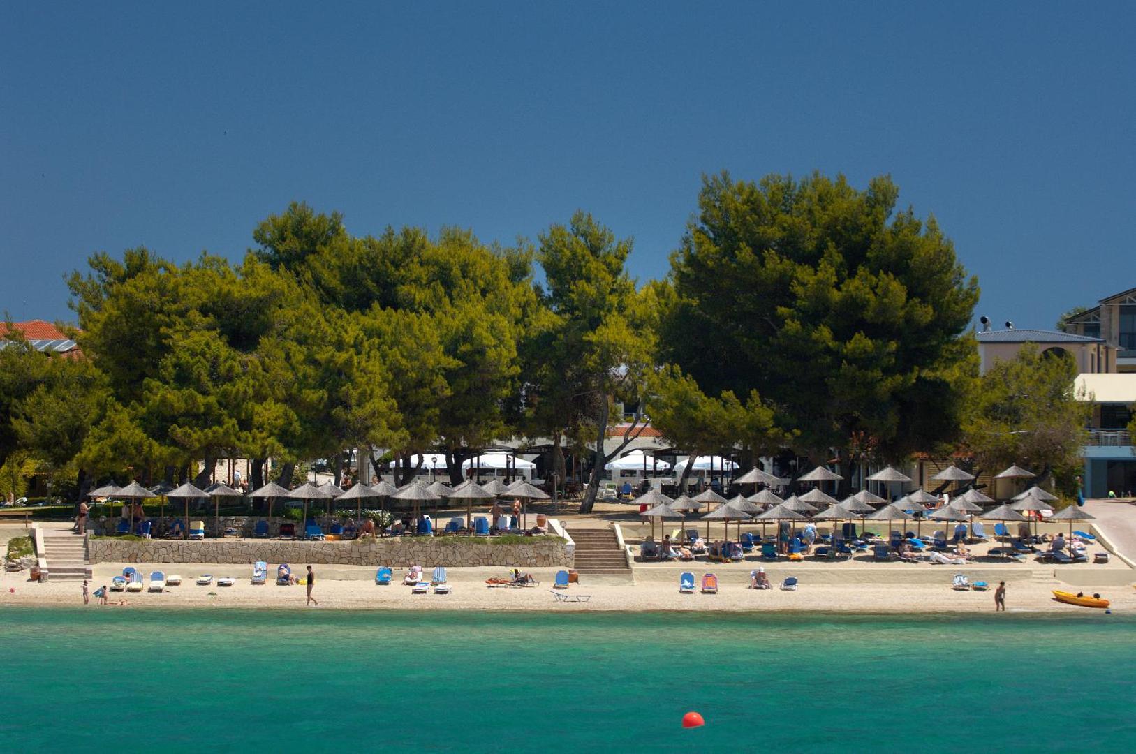 Portes Beach Hotel skats no jūras uz privāto smilšu pludmali ar saulessargiem un zaļojošiem kokiem Halkidiki piekrastē